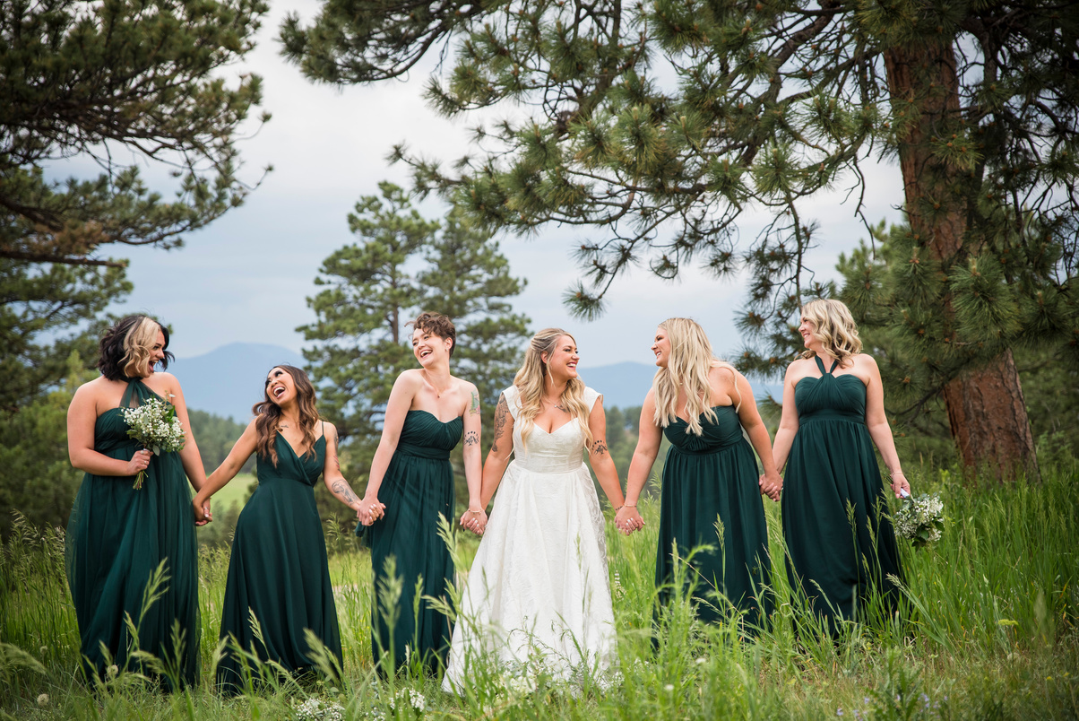 A bride and her bridesmaids hold hands and walk toward the camera, captured by Denver wedding photographer, Two One Photography.