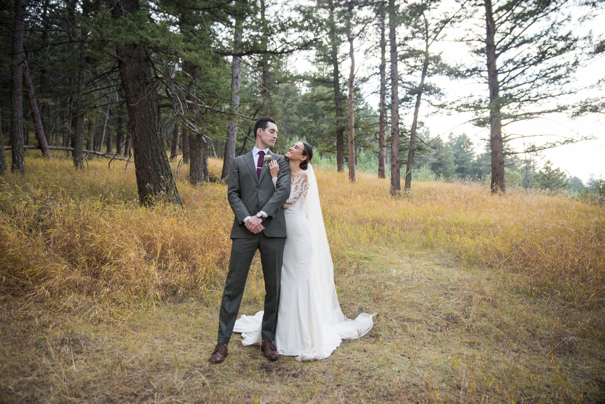 A bride embraces her groom from behind and the two smile at one another in the pine trees at The Pines at Genesee in Genesee, Colorado.