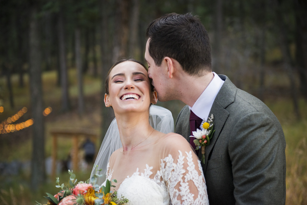 A groom kisses his bride on the cheek in the woods as she smiles at the camera.