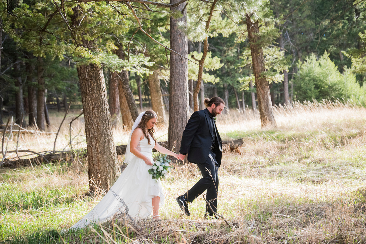 A groom casually leads his bride through the woods at The Pines at Genesee wedding venue in Denver, Colorado.